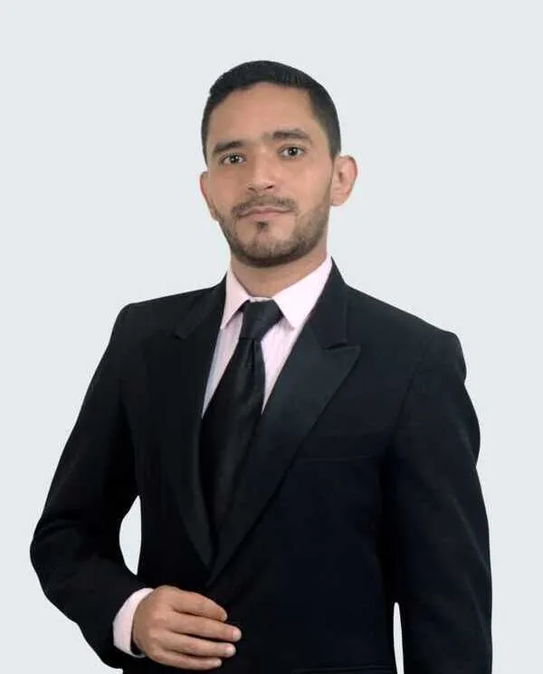 Professional portrait of a man in a black suit, light pink shirt, and black tie against a plain background.