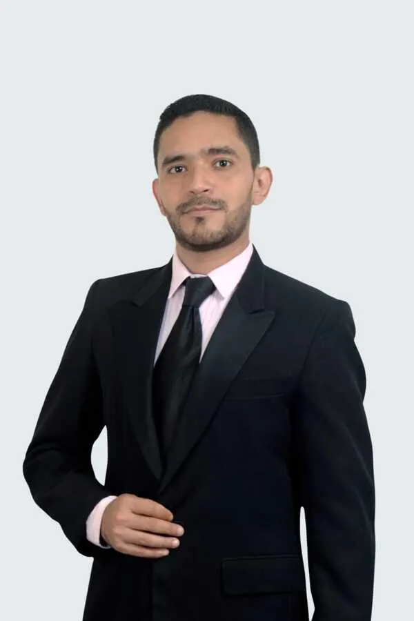 Professional portrait of a man wearing a black suit, light pink shirt, and black tie against a plain background.