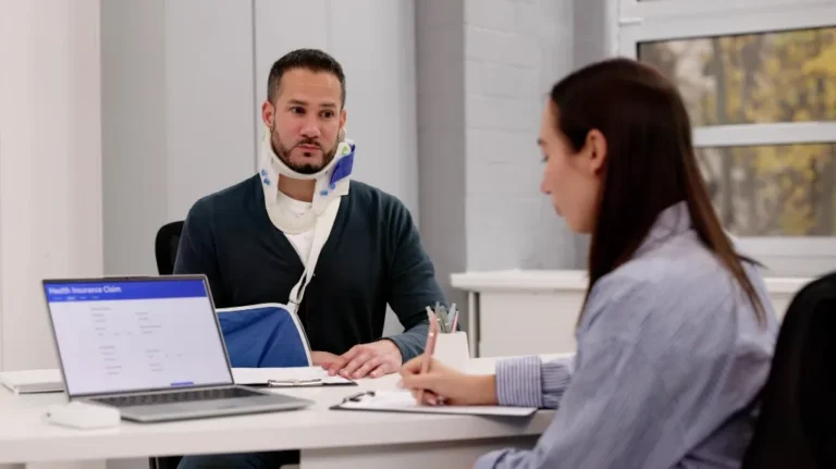 Man wearing neck brace and arm sling consulting with woman at desk, laptop displaying health insurance claim.