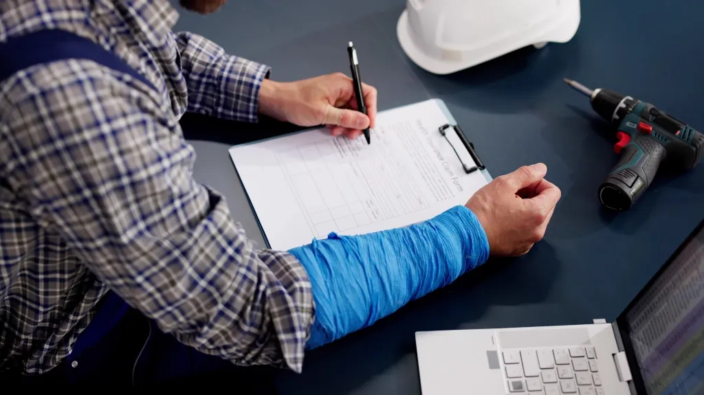 Person with a blue arm cast filling out a clipboard form on a dark desk.