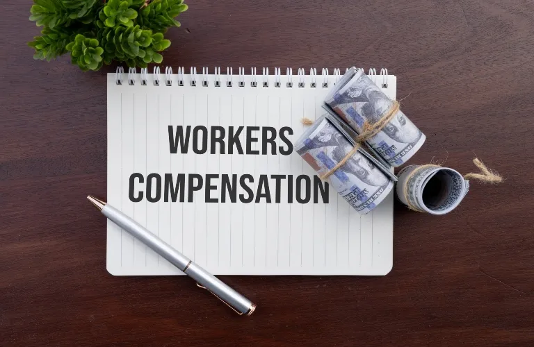 Notepad with "WORKERS COMPENSATION" text, rolled US dollar bills, silver pen, and small green plant on wooden surface.