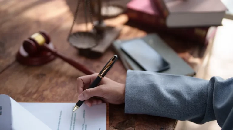 Person in a gray suit writing on a document with legal books, gavel, and scales on wooden desk.