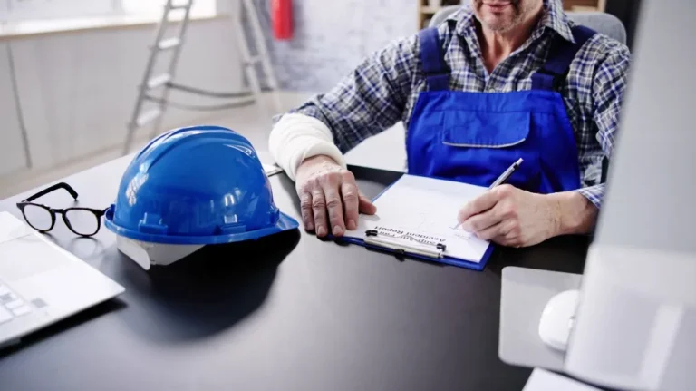 Engineer in blue overalls writing on clipboard beside blue safety helmet and eyeglasses on desk.