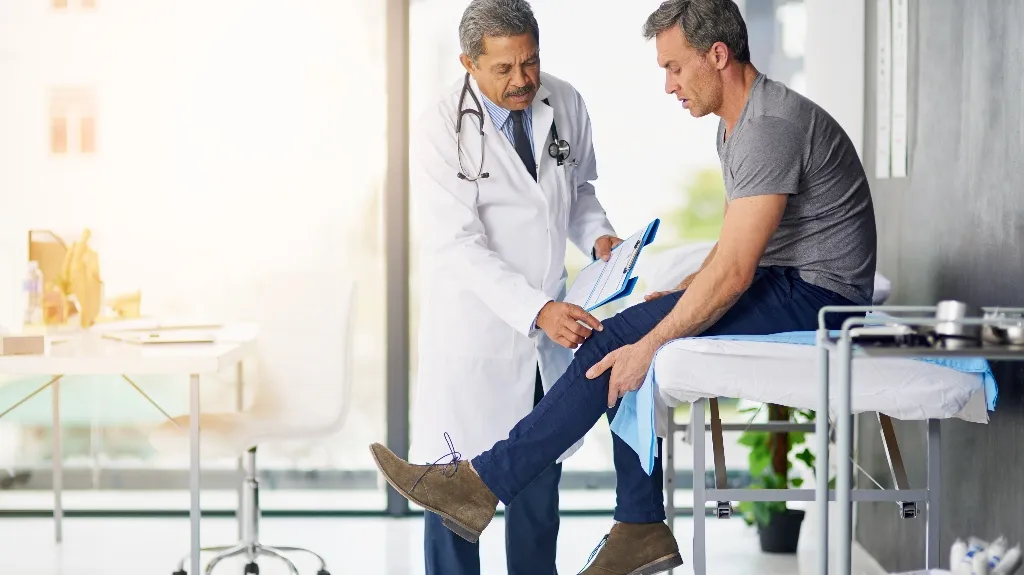 Doctor in white coat examining patient's knee in a bright medical office.