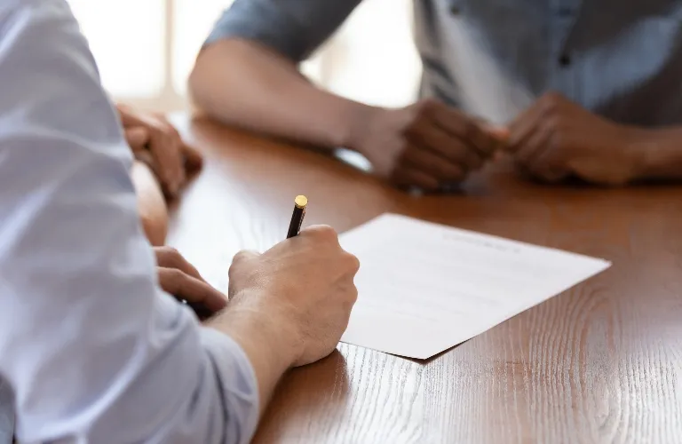 Two individuals seated at a wooden table, one signing a document with a pencil.